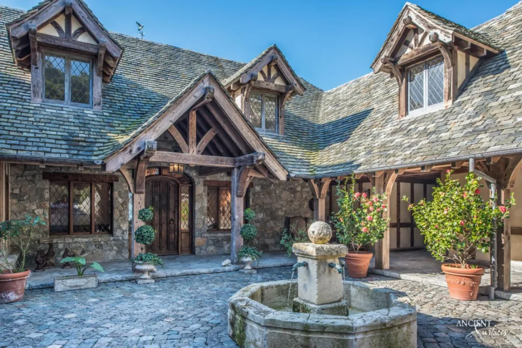 Cobblestone courtyard with a stone fountain, potted citrus trees and steep tiled gables of a stone and timber villa