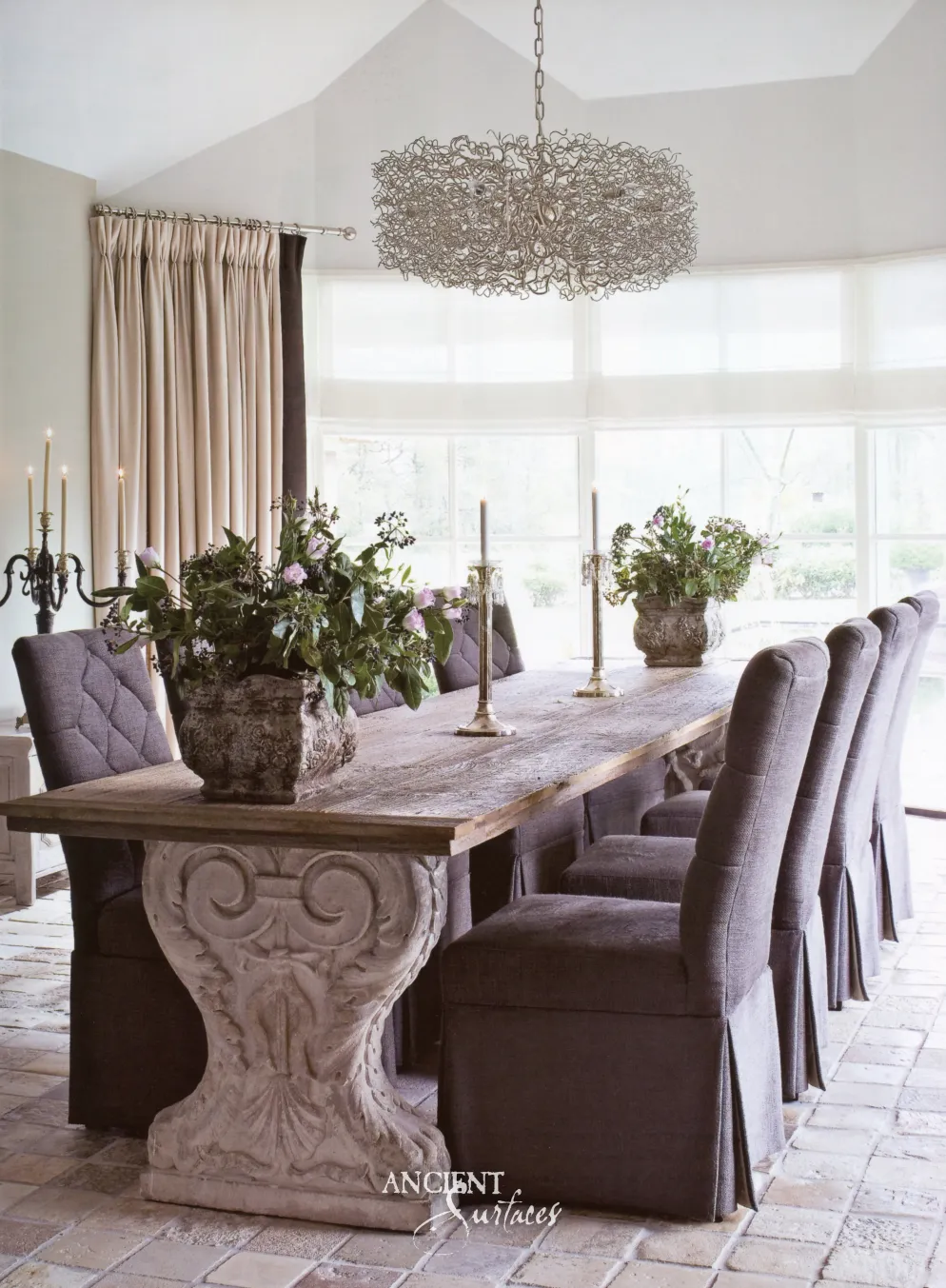Long dining table on a carved limestone pedestal base set on antique limestone floor pavers before tall garden windows