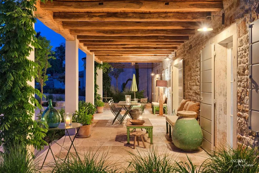 Lantern lit stone terrace lounge with wicker seating, glazed demijohns and a timber beamed pergola at dusk