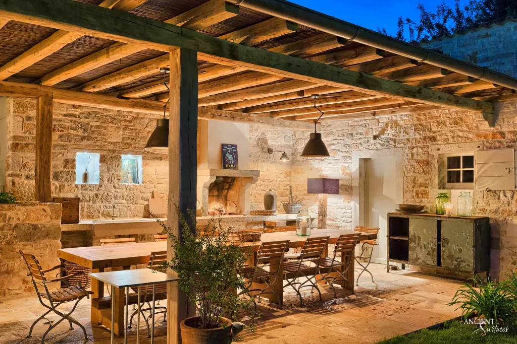 Outdoor dining terrace at dusk before a stone hearth built into a limestone clad wall