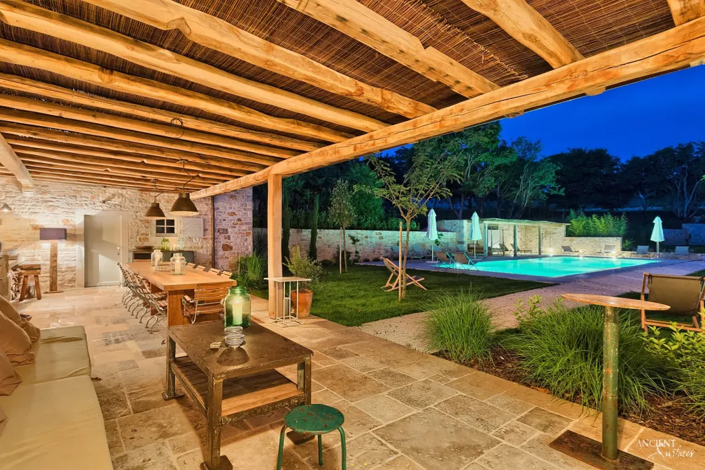Long outdoor dining table beneath a timber pergola overlooking an illuminated swimming pool at dusk