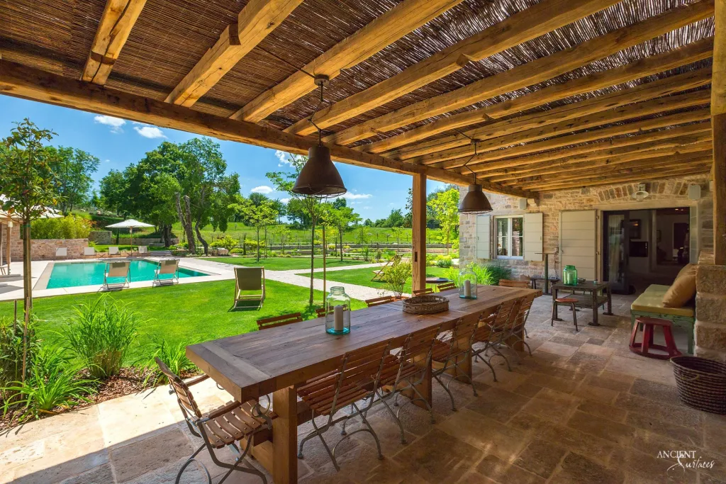 Covered stone terrace with a long dining table facing a limestone edged swimming pool and open lawn