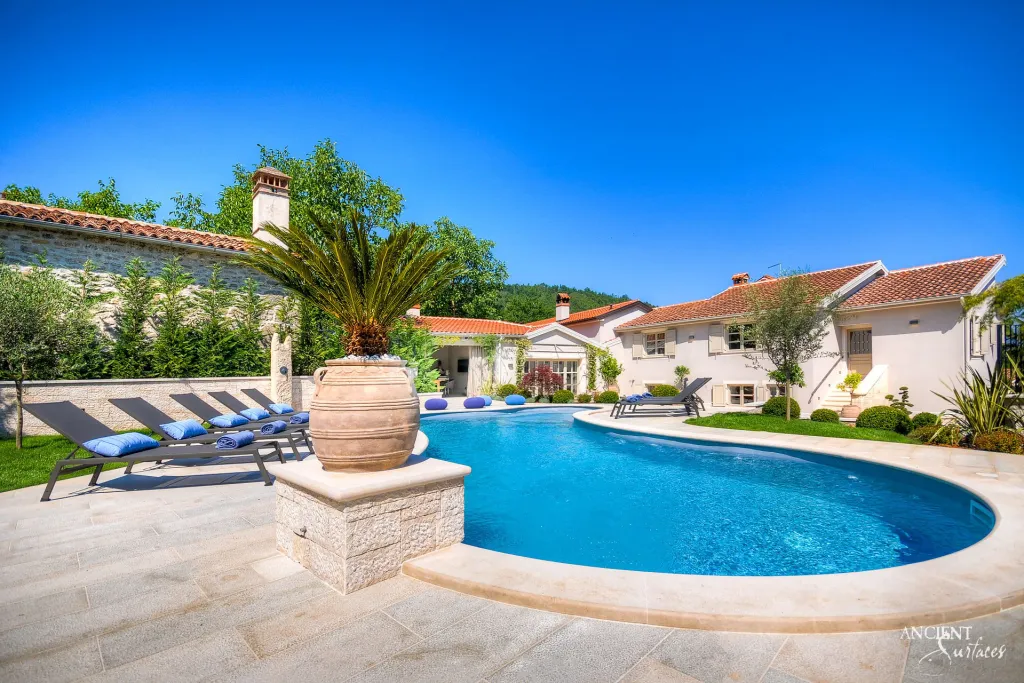 Sun lit swimming pool with pale stone coping beside a Croatian villa clad in reclaimed stone