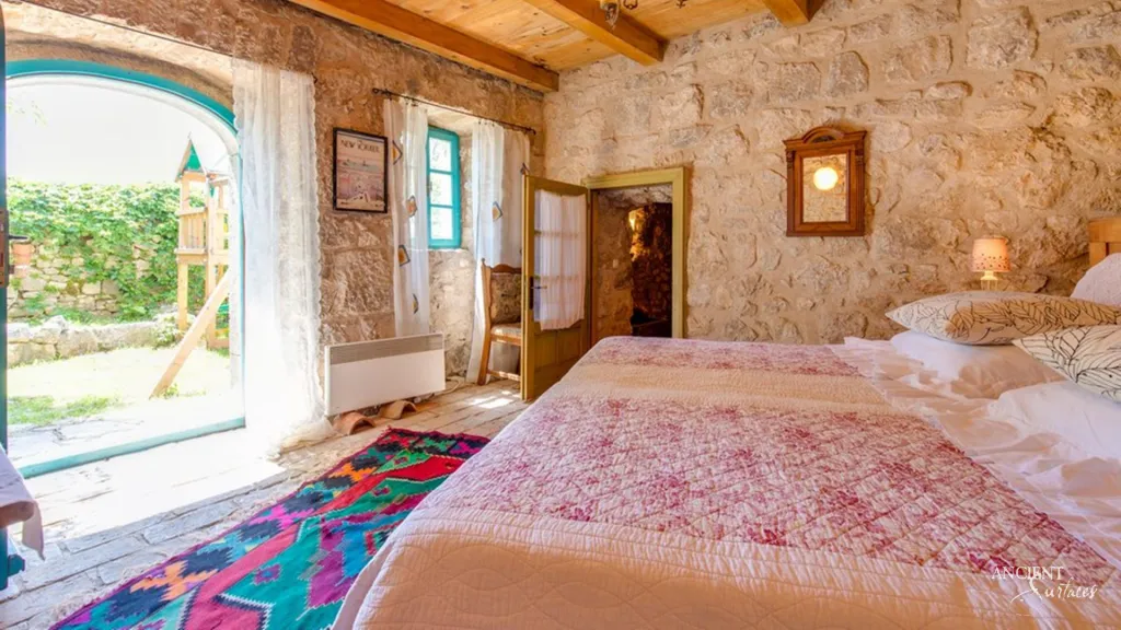 Bedroom with rubble stone walls, timber ceiling beams and an arched doorway opening to a garden